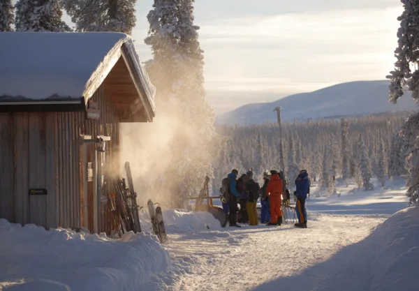 Midcent: En grupp människor njuter av en solig vinterdag vid en fjällstuga. Snötäckta träd och fjälllandskap skapar en