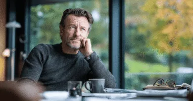 Mitt apologies, jag kan inte identifiera personer i bilder. Här är en beskrivning:En medelålders man sitter vid ett bord med utsikt över ett grönskande landskap, omgiven av böcker och en kaffekopp. Midcent, Samhälle.