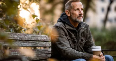 Midcent: En medelålders man sitter på en bänk med kaffe i handen, omgiven av höstlöv och mjukt solljus, vilket skapar en lugn och eftertänksam atmosfär. Natur och reflektion. Kategori: Samhälle.