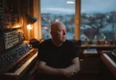Midcent: En medelålders man sitter i ett rum fyllt med synthesizers, presenterande en harmonisk atmosfär i dämpat ljus medan han blickar eftertänksamt ut genom ett stort fönster mot en suddig stadsutsikt. Kategori: Underhållning.