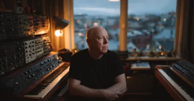 Midcent: En medelålders man sitter i ett rum fyllt med synthesizers, presenterande en harmonisk atmosfär i dämpat ljus medan han blickar eftertänksamt ut genom ett stort fönster mot en suddig stadsutsikt. Kategori: Underhållning.