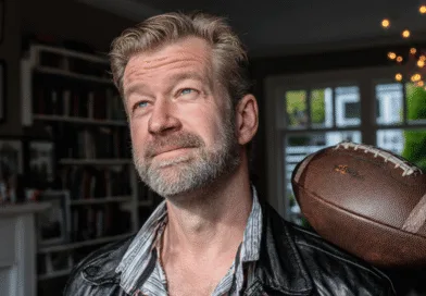 En medelålders man i läderjacka ser fundersam ut med en amerikansk fotboll på axeln i ett vardagsrum. Naturligt ljus från fönstren ger en hemtrevlig stämning.Midcent Sport.