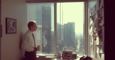 Midcent: En medelålders man står med en kaffekopp bredvid ett kontorsfönster i stadsmiljö, omgiven av papper och fotografier. Kategorin är Ekonomi.