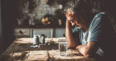 Midcent: En medelålders man sitter vid ett träbord med ett glas vatten, omgiven av dämpat ljus och en lantlig atmosfär. Fokuserad blick genom fönstret. Kategori: Samhälle.