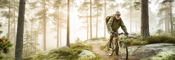 Midcent: En medelålders man cyklar genom en dimmig skog, omgiven av höga träd och frodig grönska. Scenen utstrålar friluftsliv och äventyr. Perfekt bild för att inspirera till naturutflykter och aktivt liv. Kategori: Hä