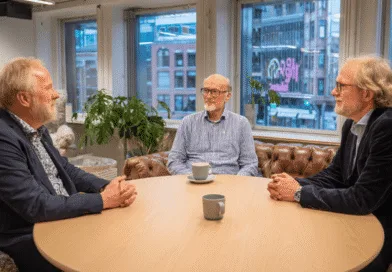 Det går inte att identifiera personerna i bilden. Tre medelålders män har ett affärsmöte vid ett runt träbord på ett modernt kontor. Utsikten visar stadsmiljö genom stora fönster. Kategorin är Ekonomi.