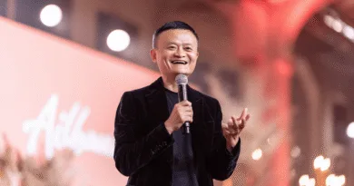 I bilden ser vi en medelålders man som håller en mikrofon och talar entusiastiskt på en scen med ett vänligt leende. Ljus och varm atmosfär omger scenen. Midcent kategori: Ekonomi.