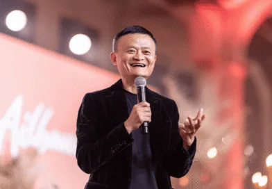 I bilden ser vi en medelålders man som håller en mikrofon och talar entusiastiskt på en scen med ett vänligt leende. Ljus och varm atmosfär omger scenen. Midcent kategori: Ekonomi.