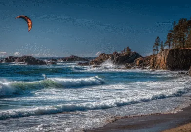 Midcent: En spektakulär kustlandskap med starka vågor och en kitesurfare mot en klarblå himmel. Perfekt för spännande vattensporter och naturlig skönhet. Kategori: Sport.