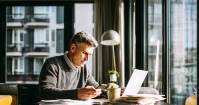 Midcent fokuserar på arbete och produktivitet i en modern miljö. En medelålders man sitter vid ett skrivbord, använder sin smartphone och laptop bland papper. Kategori: Ekonomi.