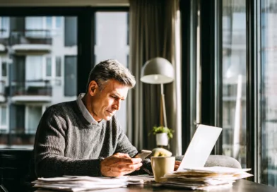 Midcent fokuserar på arbete och produktivitet i en modern miljö. En medelålders man sitter vid ett skrivbord, använder sin smartphone och laptop bland papper. Kategori: Ekonomi.