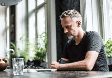 Midcent: En medelålders man sitter vid ett bord i ett ljust rum och fokuserar på en blodtrycksmätare omgiven av grönska och frukt. Hälsa.