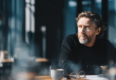 Midcent: En medelålders man i djupa tankar sitter vid ett bord med en kopp kaffe och en laptop. Ljuset ger en reflekterande och koncentrerad atmosfär. Perfekt perspektiv för inspiration och fokus. Kategori: Samhälle.