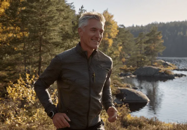 Midcent: En medelålders man joggar genom en höstfärgad skog vid en sjö, vilket skapar en känsla av frihet och hälsa. Solens strålar reflekteras i vattnet, vilket ger en inspirerande stämning. Kategori: Hälsa.
