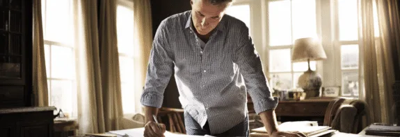 Midcent: En medelålders man fokuserar på dokument vid ett skrivbord, upplyst av naturligt ljus från stora fönster, och skapar en koncentrerad arbetsmiljö. Ekonomi.