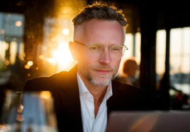 En medelålders man med glasögon arbetar fokuserat vid en laptop i solnedgången. Miljön är elegant och professionell, vilket understryker en känsla av framgång och beslutsamhet. Midcent – Ekonomi.