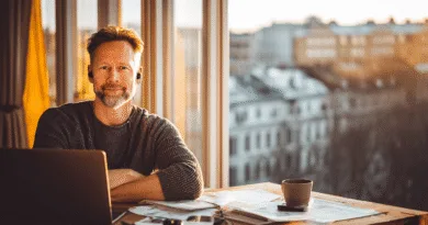 Midcent presenterar en medelålders man vid ett skrivbord med laptop, pappersarbete och kaffemugg. Solnedgång i bakgrunden genom stora fönster ger en varm och produktiv arbetsmiljö. Perfekt balans mellan arbete och avkoppling. Kategori: Samhälle.