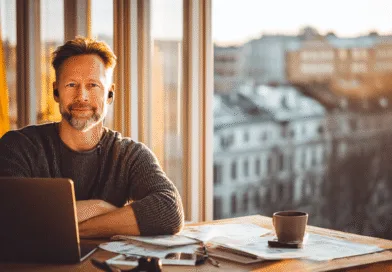 Midcent presenterar en medelålders man vid ett skrivbord med laptop, pappersarbete och kaffemugg. Solnedgång i bakgrunden genom stora fönster ger en varm och produktiv arbetsmiljö. Perfekt balans mellan arbete och avkoppling. Kategori: Samhälle.