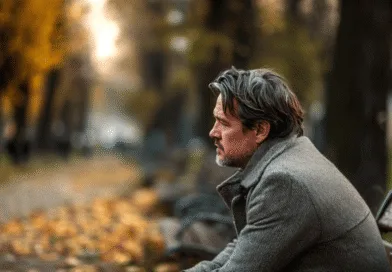 Midcent: En medelålders man sitter på en parkbänk omgiven av höstlöv, djup i tankar. En avbild av eftertänksamhet och höstlig stillhet. Samhälle.