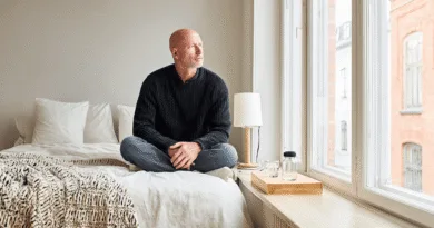 Midcent: En medelålders man sitter på en säng i ett ljust sovrum, blickar tankfullt ut genom fönstret. Minimalistisk inredning med fokus på enkelhet och komfort. Kategori: Inredning.