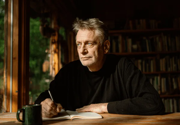 En medelålders man sitter vid ett träbord och skriver i en anteckningsbok, omgiven av böcker och med naturen utanför fönstret. Midcent. Kategori: Samhälle.