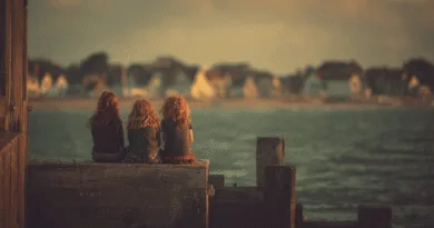 Tre barn sitter på en brygga och blickar ut över dimmiga hus vid vattnet i solnedgången. Midcent Resa.