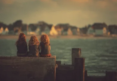 Tre barn sitter på en brygga och blickar ut över dimmiga hus vid vattnet i solnedgången. Midcent Resa.