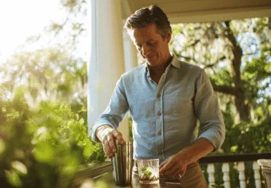 Midcent: En medelålders man står på en veranda och blandar en drink i en cocktailshaker. Solen skiner i bakgrunden och omgivningen är lummig och grönskande. En avkopplande atmosfär genomsyrar bilden. Kategori: Mat.