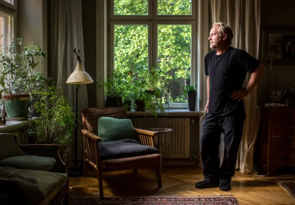 Midcent: En vuxen man står i ett ljust rum omgiven av växter, vilket skapar en lugn atmosfär. Kategorin är Samhälle.