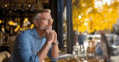 En medelålders man sitter vid ett kafébord och blickar ut genom fönstret, djup i tankar. Höstlöven i bakgrunden skapar en lugn atmosfär. Midcent. Samhälle.