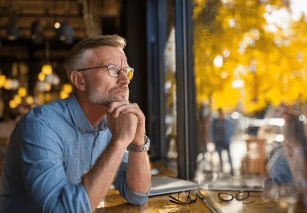 En medelålders man sitter vid ett kafébord och blickar ut genom fönstret, djup i tankar. Höstlöven i bakgrunden skapar en lugn atmosfär. Midcent. Samhälle.