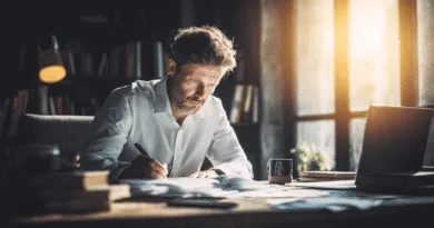 Midcent: En medelålders man fokuserad på arbete. Han sitter vid ett skrivbord i ett solbelyst rum med en laptop och dokument. Atmosfären är kreativ och inspirerande, ideal för produktivitet. Kategori: Samhälle.