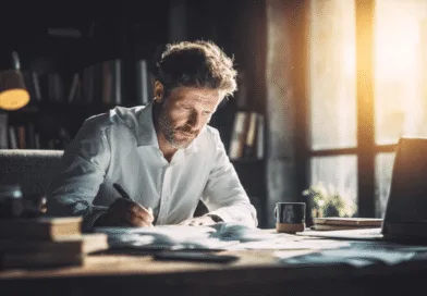 Midcent: En medelålders man fokuserad på arbete. Han sitter vid ett skrivbord i ett solbelyst rum med en laptop och dokument. Atmosfären är kreativ och inspirerande, ideal för produktivitet. Kategori: Samhälle.