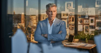 En medelålders man i en modern kontorsmiljö med armarna i kors och bestämd blick. Bakgrunden visar ett kreativt arbetsutrymme med foton och dokument. Midcent. Kategori: Ekonomi.