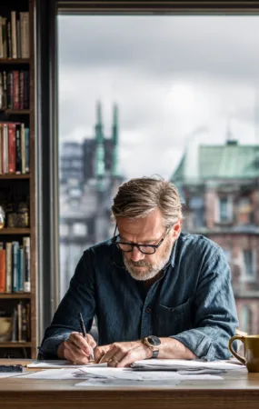 Midcent – En vuxen man arbetar fokuserat vid ett skrivbord med böcker i bakgrunden. Kreativitet och produktivitet i en inspirerande miljö. Kategori: Samhälle.