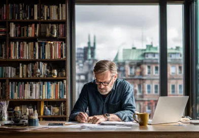 Midcent – En vuxen man arbetar fokuserat vid ett skrivbord med böcker i bakgrunden. Kreativitet och produktivitet i en inspirerande miljö. Kategori: Samhälle.