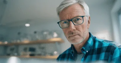 En medelålders man med grått hår och glasögon. Han ser fokuserad ut i en modern miljö med hyllor i bakgrunden. Bilden kan associeras med teknologi eller arbetsmiljö. Midcent. Kategori: Teknik.