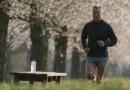 Midcent: En medelålders man joggar i en grönskande park med blommande träd. En vattenflaska står på en bänk i förgrunden. Perfekt bild av hälsa, kondition och utomhusaktiviteter. Kategori: Hälsa.