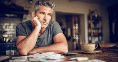 En medelålders man sitter tankfullt vid ett bord, omgivet av papper och en miniräknare, vilket tyder på ekonomiska överväganden. Midcent, Ekonomi.