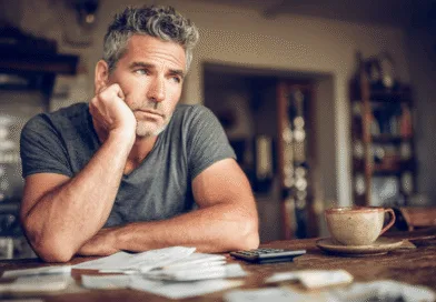 En medelålders man sitter tankfullt vid ett bord, omgivet av papper och en miniräknare, vilket tyder på ekonomiska överväganden. Midcent, Ekonomi.