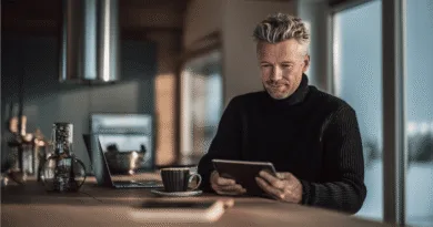 Midcent: En medelålders man i en modern miljö med laptop och surfplatta. Fokus på teknik och digital interaktion. Naturligt ljus från stora fönster ger en avslappnad atmosfär för arbetet. Kategori: Teknik.