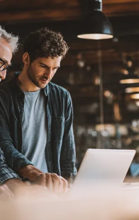 Midcent: Två män samarbetar vid en dator i ett modernt kontorsmiljö. Den ena är en medelålders man med skägg och glasögon.