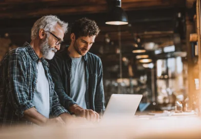 Midcent: Två män samarbetar vid en dator i ett modernt kontorsmiljö. Den ena är en medelålders man med skägg och glasögon.
