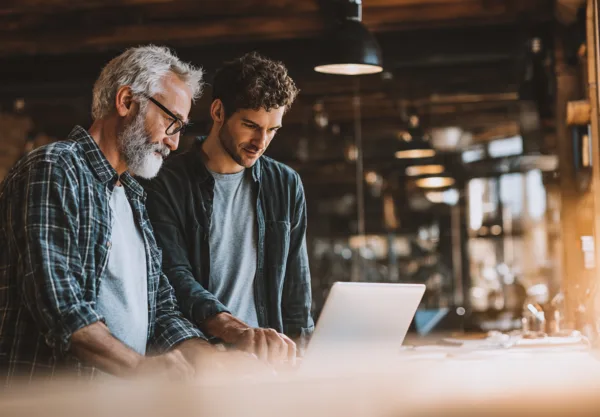Midcent: Två män samarbetar vid en dator i ett modernt kontorsmiljö. Den ena är en medelålders man med skägg och glasögon.