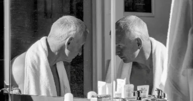 Midcent: En medelålders man tittar i spegeln i ett badrum, omgiven av hudvårdsprodukter, iklädd en handduk. Bilden fångar en stund av personlig reflektion och egenvård. Hälsa.