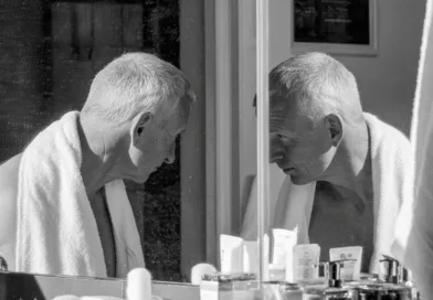 Midcent: En medelålders man tittar i spegeln i ett badrum, omgiven av hudvårdsprodukter, iklädd en handduk. Bilden fångar en stund av personlig reflektion och egenvård. Hälsa.