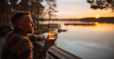 En medelålders man njuter av en solnedgång vid sjön, med ett glas whiskey i handen på en verandabrygga, omgiven av naturens lugn. Midcent Resa.