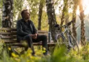 Midcent visar en medelålders man som sitter på en bänk i naturen, omgiven av gröna träd och cyklar. Denna scen förmedlar lugn och avkoppling, perfekt för friluftsentusiaster.Kategori: Samhälle.