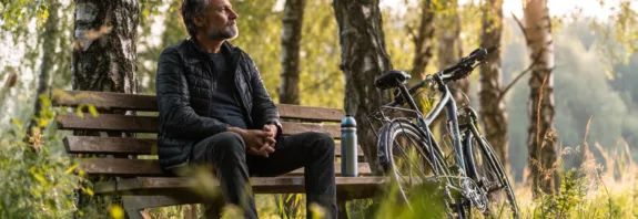 Midcent visar en medelålders man som sitter på en bänk i naturen, omgiven av gröna träd och cyklar. Denna scen förmedlar lugn och avkoppling, perfekt för friluftsentusiaster.Kategori: Samhälle.