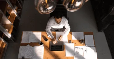 Midcent: En medelålders man jobbar fokuserat vid ett skrivbord täckt med papper och en laptop, omgiven av bokhyllor. Perfekt belysning skapar en koncentrerad atmosfär. Ekonomi.